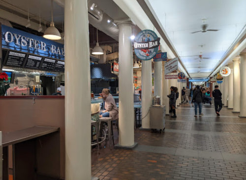 A bustling food market with various stalls, featuring an oyster bar and people dining and walking through the space.