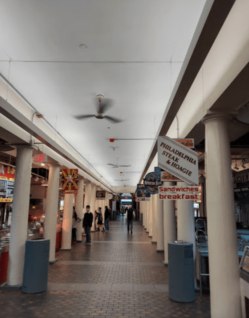 A bustling market corridor with food stalls, signs for sandwiches, and ceiling fans, featuring people walking through.