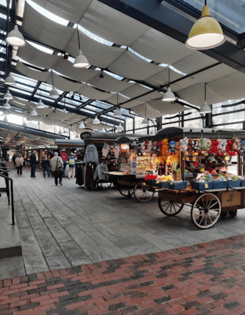A bustling market with colorful stalls, carts, and shoppers under a covered roof. Bright lights illuminate the space.