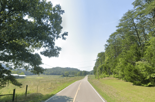 A scenic rural road lined with trees, leading into a distant hilly landscape under a clear blue sky.