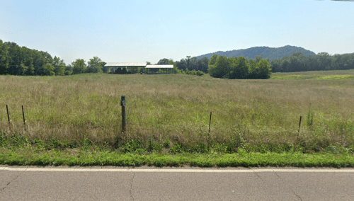 A grassy field with a distant barn and trees, set against a backdrop of rolling hills under a clear blue sky.