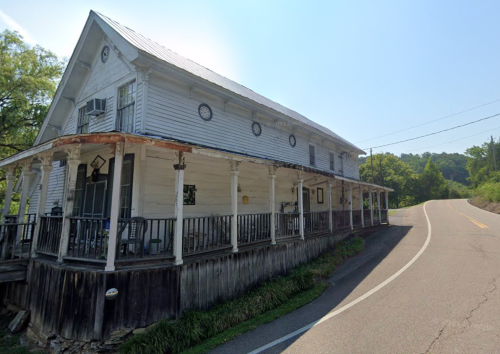 A weathered two-story house with a porch, situated beside a winding road under a clear blue sky.