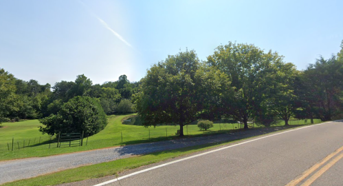 A peaceful rural scene featuring a road alongside a grassy area with trees and a pond in the background under a clear sky.