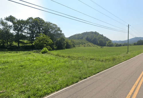A rural landscape featuring a grassy field, trees, and a distant hill under a clear blue sky. Power lines run along the road.