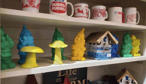Colorful garden gnomes and a yellow mushroom figurine displayed on a shelf with various mugs and signs.