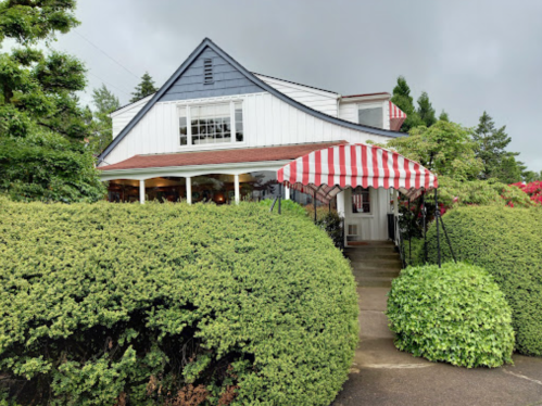 A charming house with a red and white striped awning, surrounded by lush green bushes and trees under a cloudy sky.