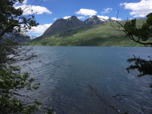 A serene lake surrounded by lush greenery and mountains under a blue sky with scattered clouds.