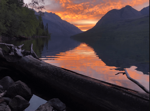 A serene lake at sunset, reflecting vibrant orange and purple hues, surrounded by mountains and lush greenery.