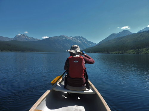 A person paddles a canoe on a calm lake, surrounded by mountains and clear blue skies.