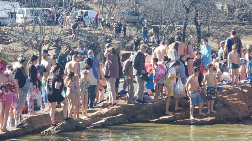 A crowded rocky area by a lake, with people in swimsuits enjoying a sunny day. Trees and vehicles are in the background.
