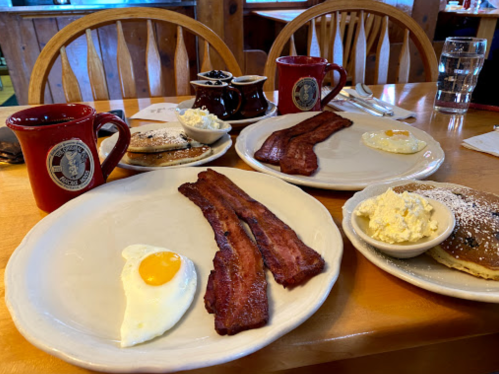 A cozy breakfast table with two plates of bacon, eggs, pancakes, and coffee mugs, set in a rustic dining area.