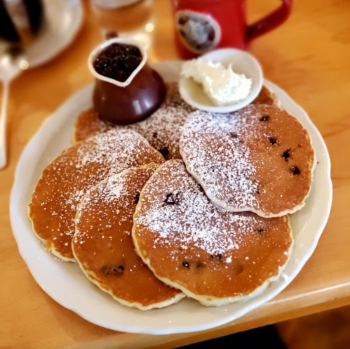 A plate of five fluffy pancakes dusted with powdered sugar, served with a small dish of chocolate sauce and whipped cream.