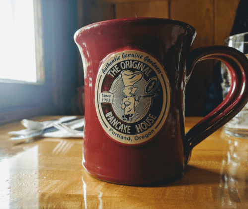 A red coffee mug with a logo for "The Original Pancake House" from Portland, Oregon, on a wooden table.