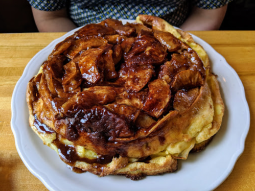 A large, fluffy pancake topped with caramelized apples on a white plate, served on a wooden table.