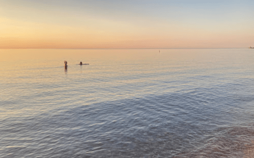 Two people wade in calm water at sunset, with soft pastel colors reflecting on the surface.