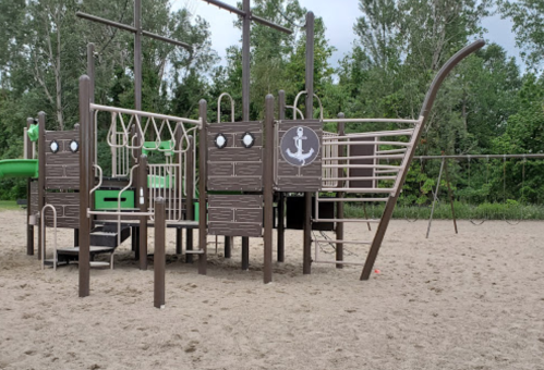 Playground structure resembling a ship, with slides, climbing features, and swings, set in a sandy area surrounded by trees.