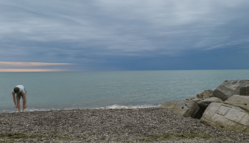A person bends down near the water's edge on a rocky beach under a cloudy sky.