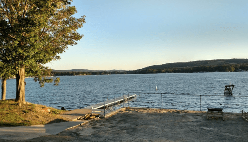 A serene lakeside view with a dock, trees, and distant hills under a clear sky.