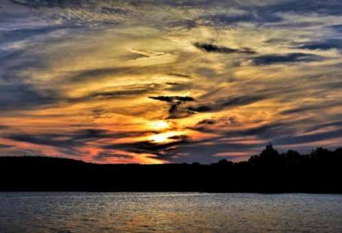 A vibrant sunset over a calm lake, with swirling clouds in shades of orange, yellow, and blue reflecting on the water.