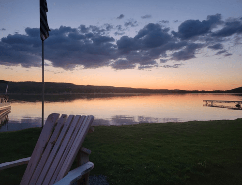 A serene lakeside view at sunset, with clouds reflecting on the water and a wooden chair in the foreground.