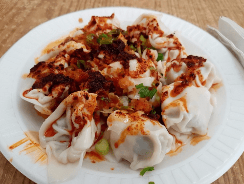 A plate of dumplings topped with spices, green onions, and a savory sauce, served on a paper plate.