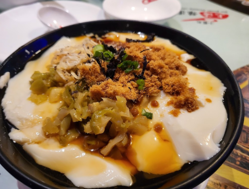 A bowl of silky tofu topped with shredded chicken, pickled vegetables, and a sprinkle of seasoning.