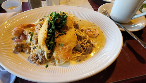 A plate of creamy pasta topped with fried fish, shrimp, and fresh greens, served with a side of sauce.