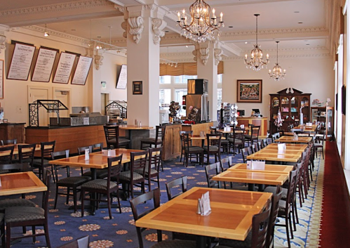 A spacious restaurant interior with wooden tables, chairs, chandeliers, and a menu board on the wall.