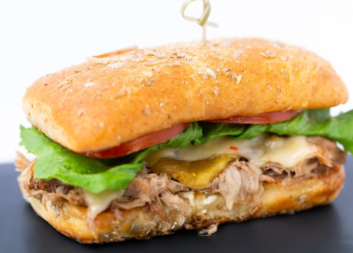 A close-up of a sandwich with pulled pork, lettuce, tomato, pickles, and melted cheese on a toasted bun.