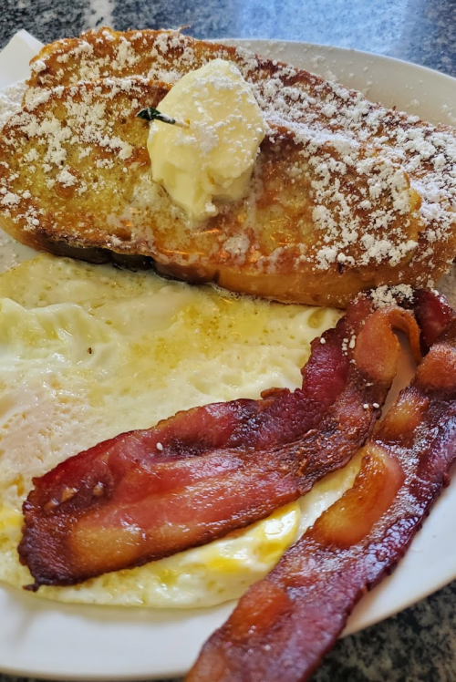 A plate of French toast topped with butter and powdered sugar, served with eggs and crispy bacon.