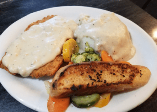 A plate of fried chicken with creamy gravy, mashed potatoes, and mixed vegetables, alongside a toasted bread slice.
