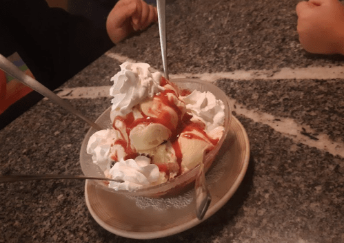 A bowl of ice cream topped with whipped cream and strawberry sauce, served with three spoons on a marble table.