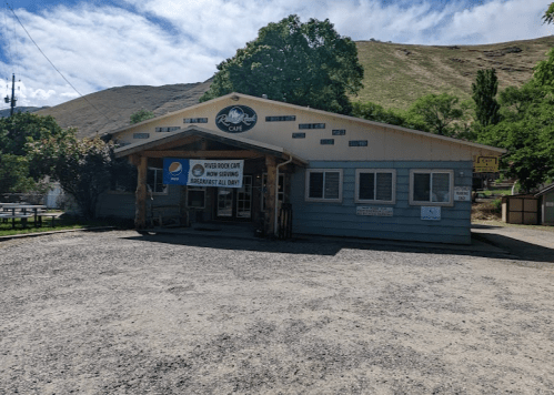 A rustic café building with a gravel parking area, surrounded by trees and hills in the background.