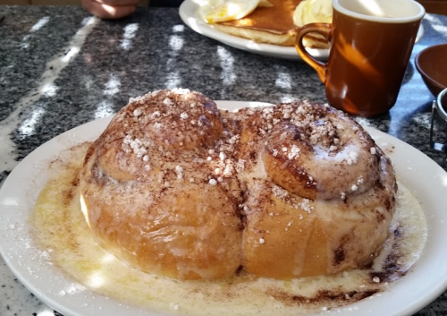 A large cinnamon roll drizzled with icing and sprinkled with powdered sugar, served on a white plate.