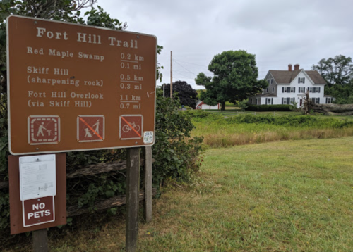 Sign for Fort Hill Trail with directions and distances, overlooking a grassy area and a large house in the background.