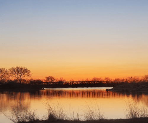 A serene sunset over a calm river, with silhouettes of trees and a bridge against a colorful sky.