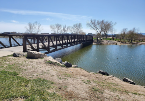 A metal bridge spans a calm lake, surrounded by grassy banks and bare trees under a clear blue sky.