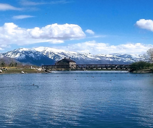 A serene lake with a wooden bridge, surrounded by snow-capped mountains and a clear blue sky with fluffy clouds.