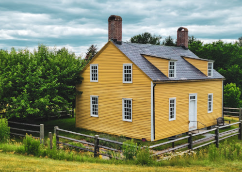 A yellow two-story house with a gray roof, surrounded by greenery and a wooden fence under a cloudy sky.