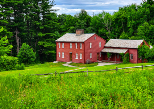 A red two-story house surrounded by lush green grass and trees under a cloudy sky.