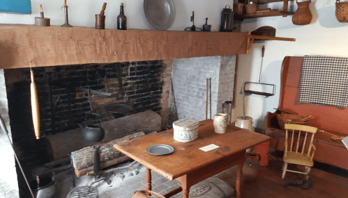 A rustic kitchen with a large fireplace, wooden table, chairs, and various vintage kitchen tools and utensils displayed.