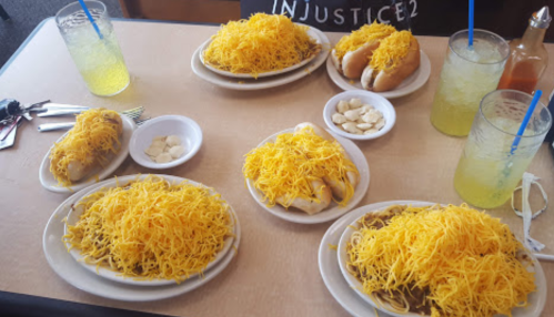 A table filled with plates of chili cheese spaghetti, hot dogs topped with cheese, and drinks.