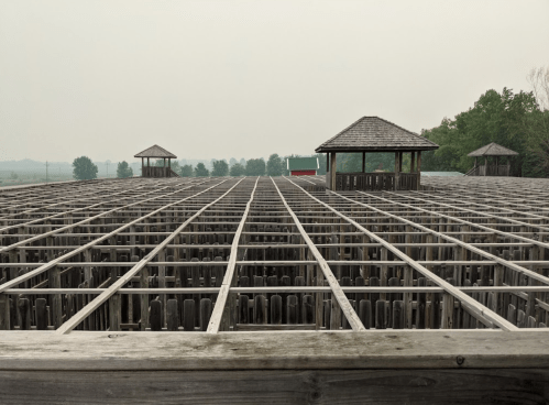 A wooden structure with a grid pattern, surrounded by trees and a hazy sky, featuring two small pavilions.