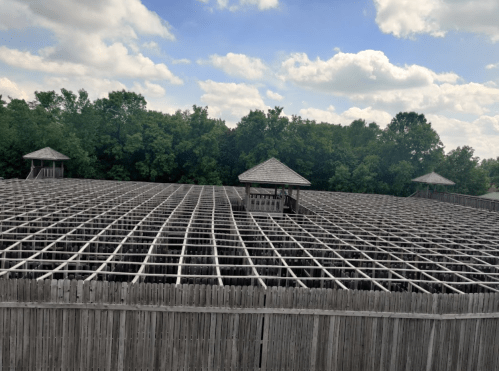 A large wooden structure with a grid pattern, surrounded by trees and under a cloudy sky.