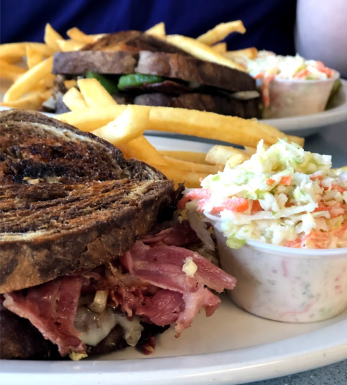 A close-up of a sandwich with corned beef, served with fries and a side of coleslaw on a plate.
