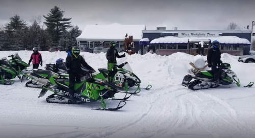A group of snowmobilers in winter gear prepares to ride in a snowy area near a building with a blue awning.