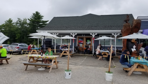 Outdoor seating area of a restaurant with umbrellas, people dining, and a wooden sculpture nearby on a cloudy day.