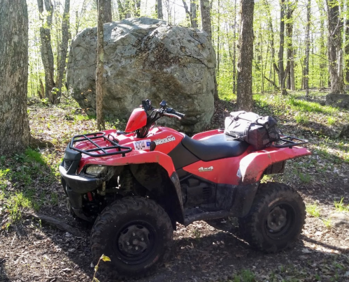 A red ATV parked near a large rock in a wooded area with green trees and sunlight filtering through.