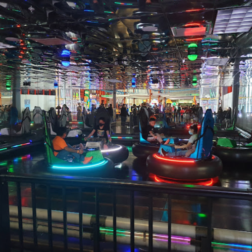 Four children in colorful bumper cars inside a vibrant arcade, with bright lights and a lively atmosphere.