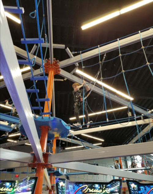A person navigates an indoor ropes course, suspended high above the ground, surrounded by colorful structures and lights.